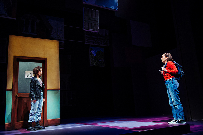 Kristen DiMercurio as Joan in Fun Home, opposite Sarah Masterson as Medium Alison; Kristen stands in front of an office door with a computer printed sign that says GAY UNION on it.