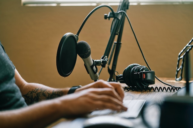 A pair of tattooed hands working at a keyboard next to a microphone with a pop filter and a pair of headphones.