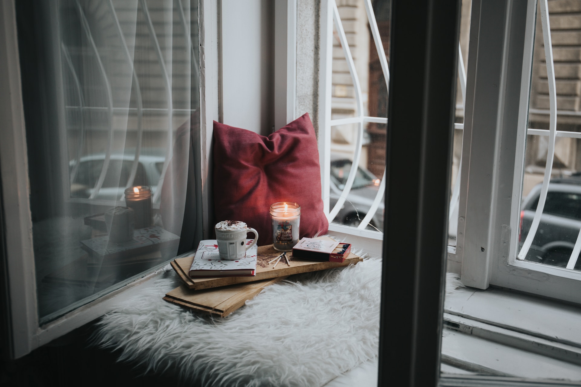 A cozy set-up in a window bench covered with a shaggy rug, with a candle, a pillow, a mug, a journal, and a box of matches.