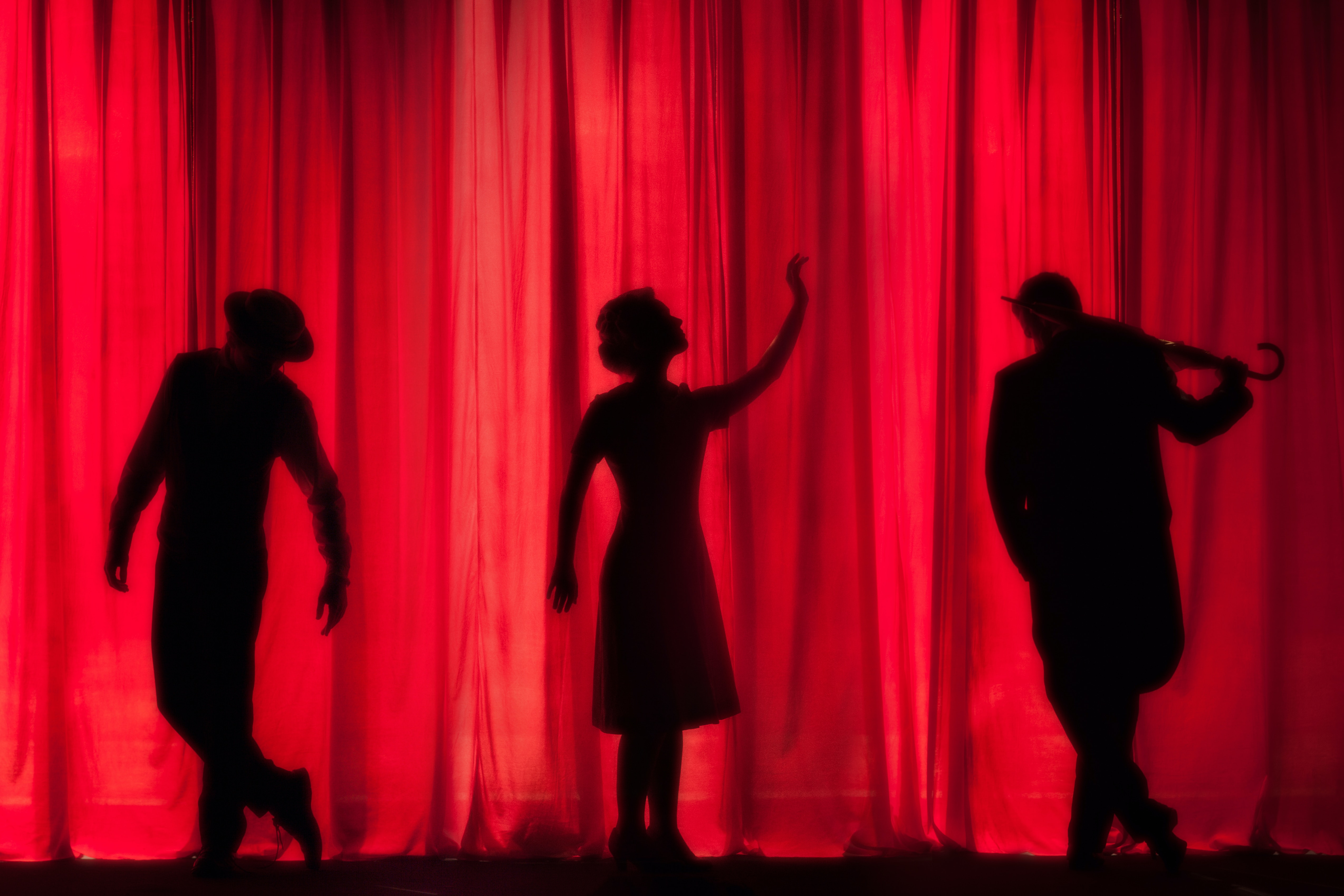 Three silhouettes behind a red theatre curtain, of a jaunty man in a flat cap, a woman in a dress gesticulating dramatically upward, and someone holding a fiddle.