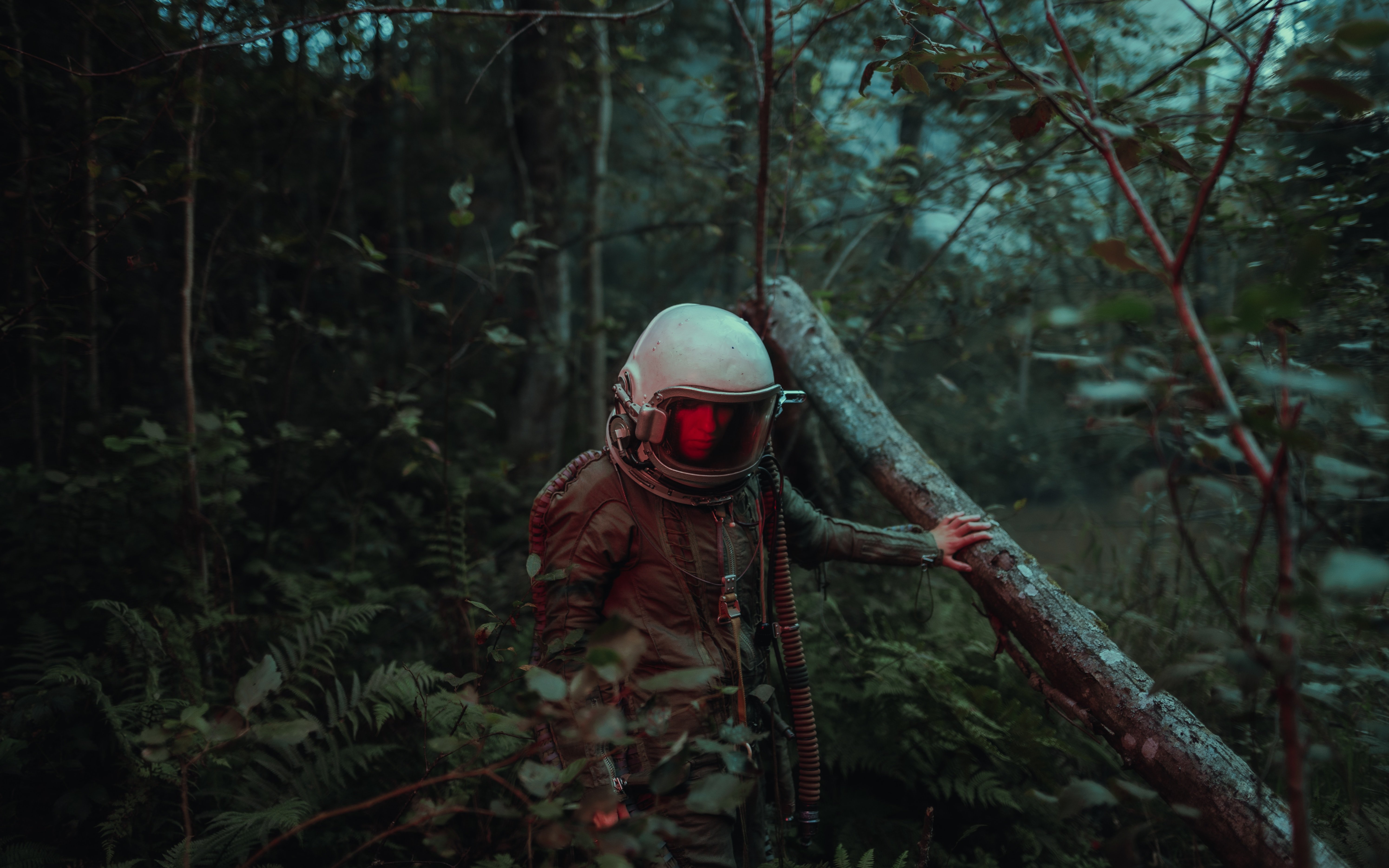 An astronaut in a dirty, old suit and a red tinted visor, walking through a dark forest, holding onto a tumbled tree.